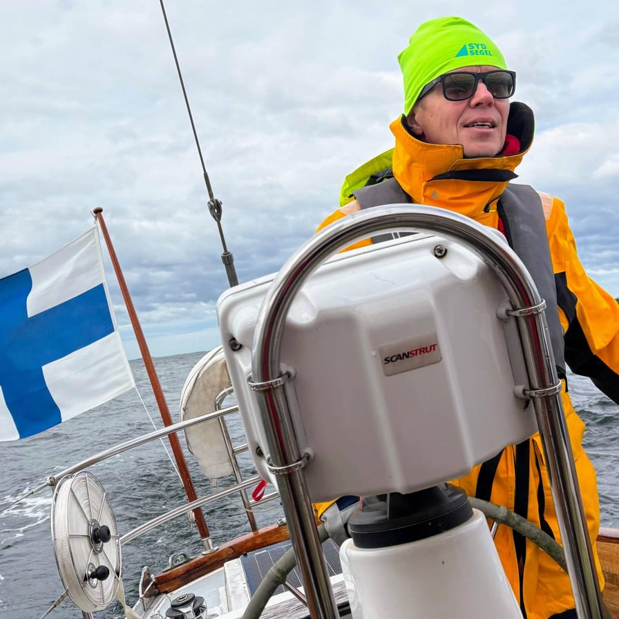 Kuva Harri Kiljanderista. Hän seisoo purjeveneen kannella aurinkolasit ja neonvihreä pipo päässään, yllään keltainen purjehdustakki ja pelastusliivi.