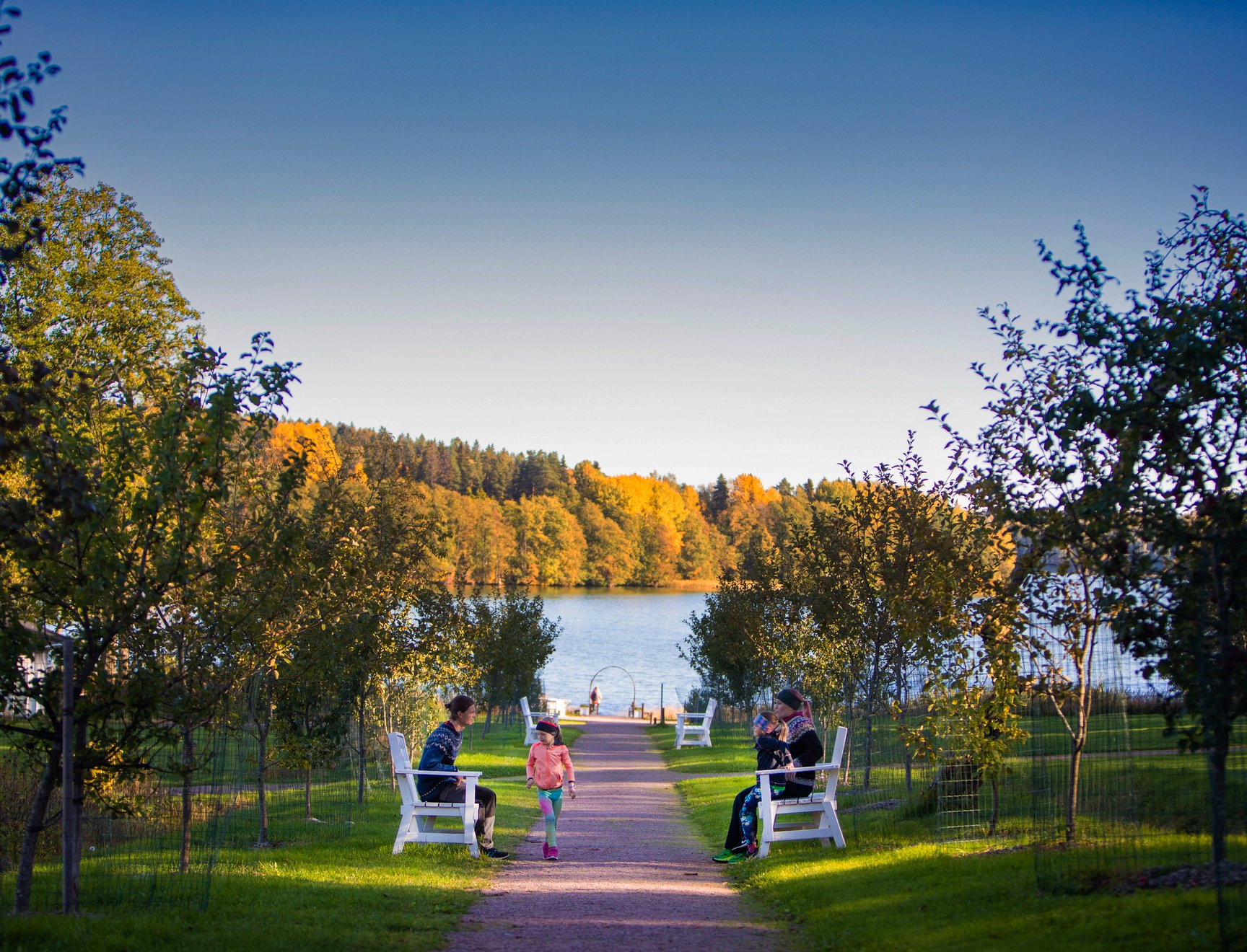 Kuva Wohls Gårdin rannalta. Kuvassa hiekkatie rannalle, jonka varrella muutamia valkoisia penkkejä. Kahdella penkillä istuu ihmisiä, hiekkatiellä kävelee lapsi.