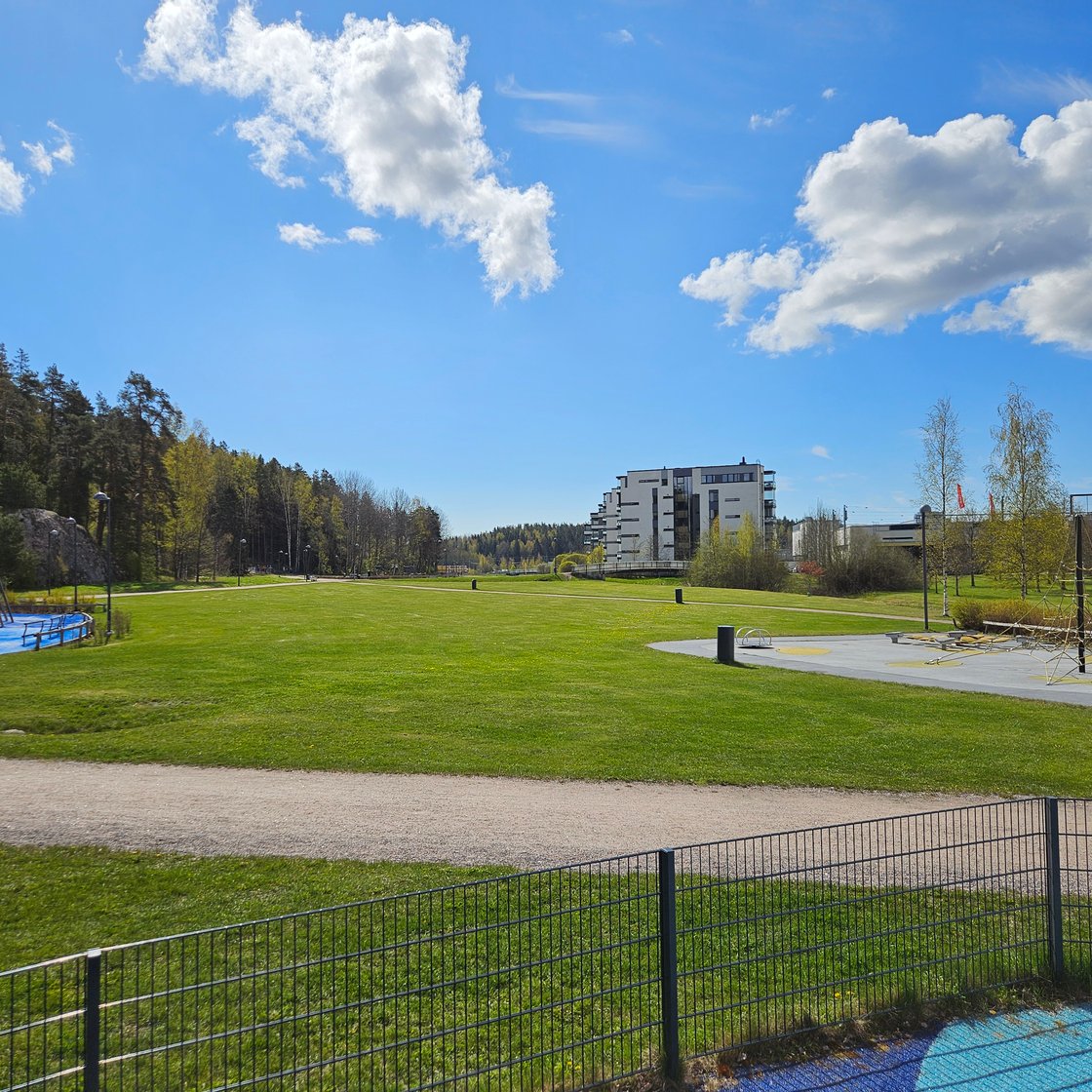 Kirkkonummi Kirsikkapuisto park on a sunny summer day. Mostly lawn in view, apartment buildings in the background and some playground equipment in the foreground