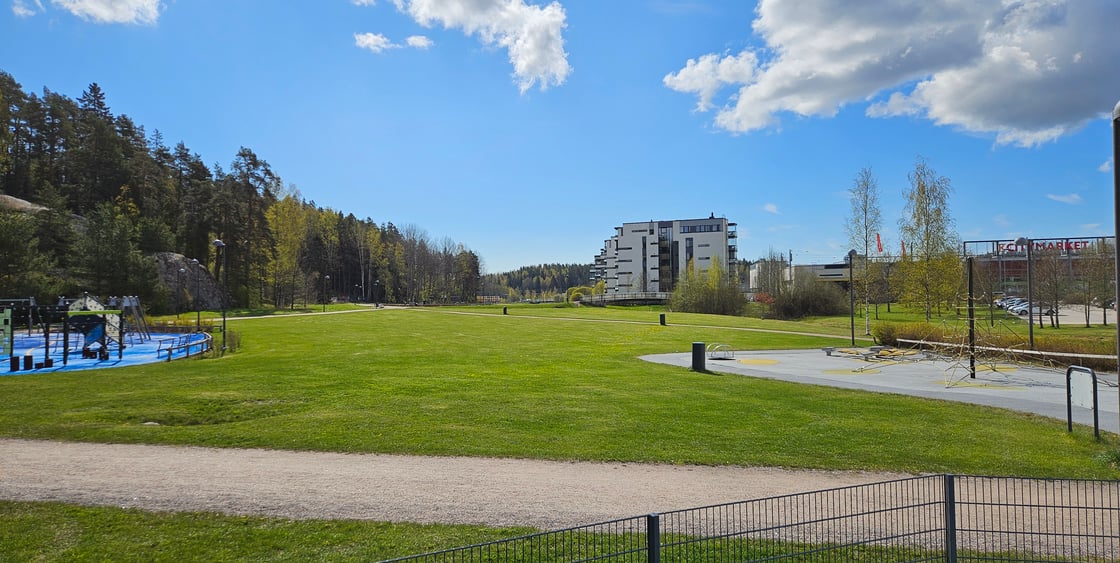 Kirkkonummen Kirsikkapuisto aurinkoisena kesäpäivänä. Näköpiirissä pääasiassa nurmialuetta, taustalla kerrostaloja ja edustalla hieman leikkipuistoja