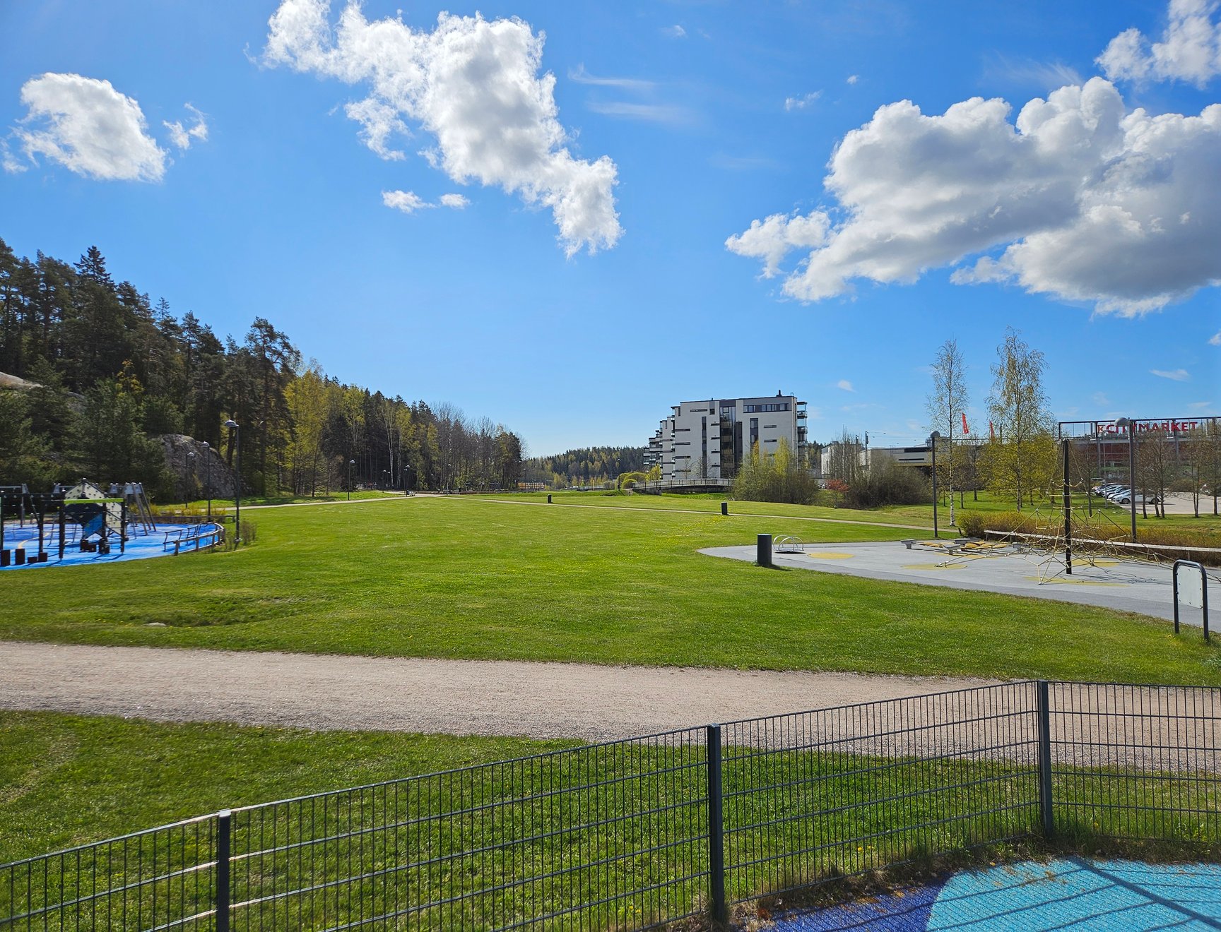 Kyrkslätt Kirsikkapuisto en solig sommardag. Nästan bara gräsmatta i blickfånget, i bakgrunden flervåningshus och i förgrunden lite lekplatsutrustning