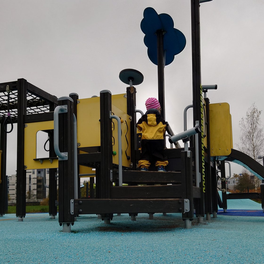 A small child in overalls climbing on a play structure with a slide and various climbing frames.