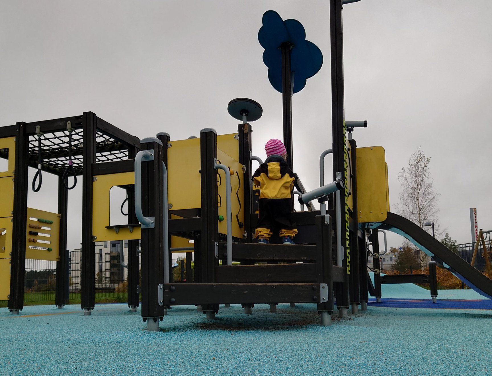 A small child in overalls climbing on a play structure with a slide and various climbing frames.