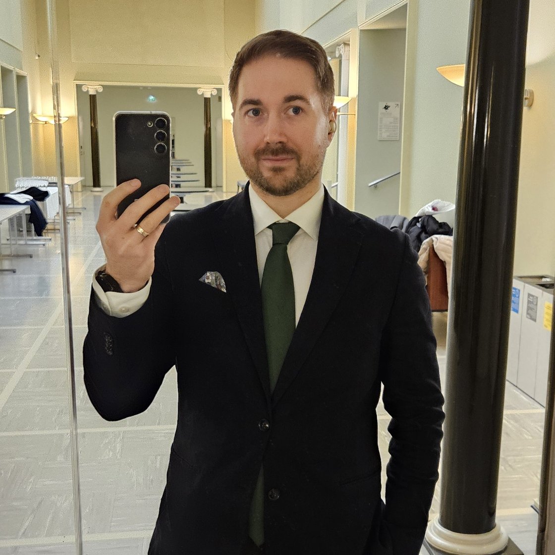 Lauri Lavanti standing in front of a mirror with a phone in his hand, taking a selfie. Wearing a black jacket, white shirt and green tie. In the background, the light corridor of the main building of the University of Helsinki.