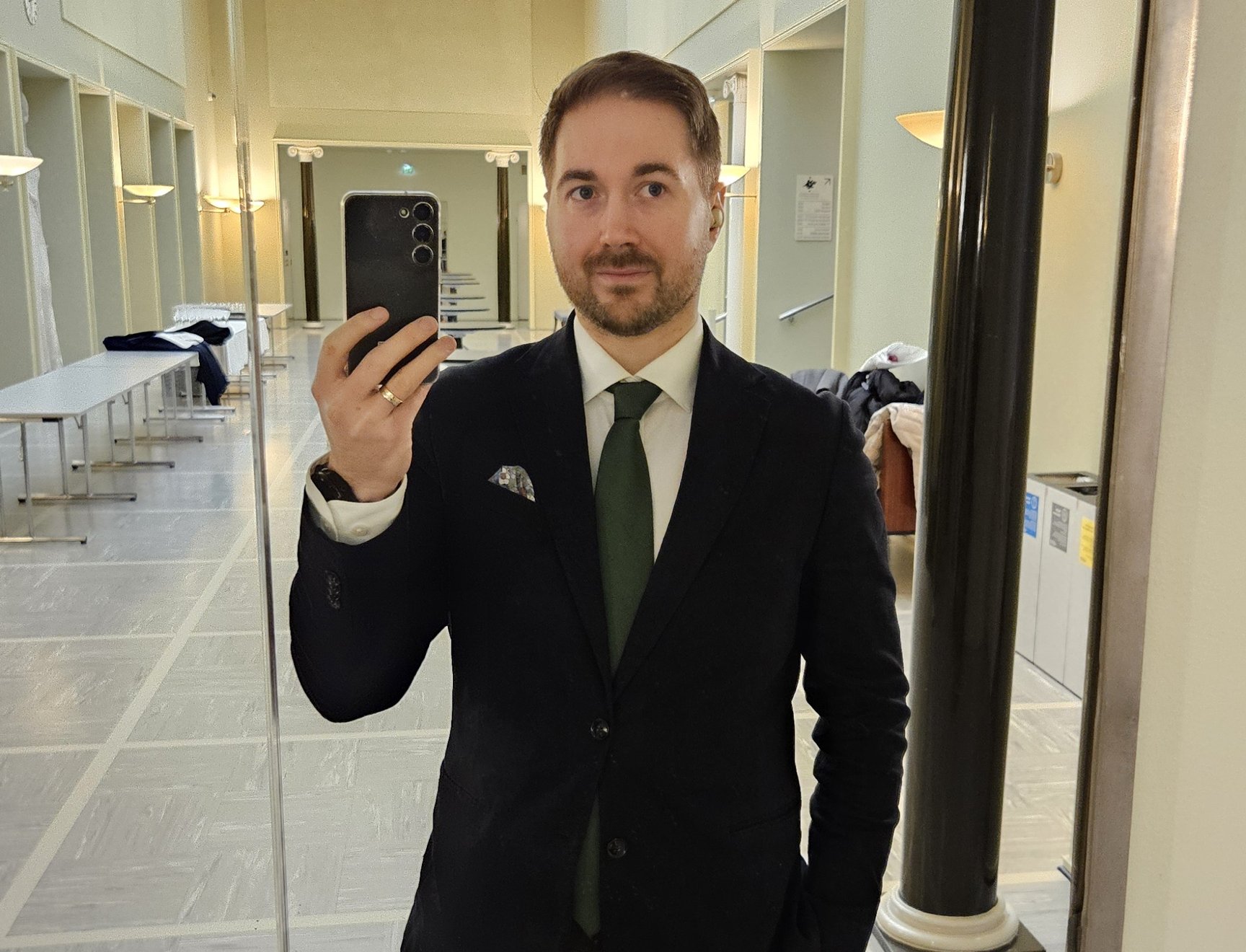 Lauri Lavanti stående framför en spegel med en telefon i handen och tar en selfie. Klädd i svart jacka, vit skjorta och grön slips. I bakgrunden den ljusa korridoren i Helsingfors universitets huvudbyggnad.