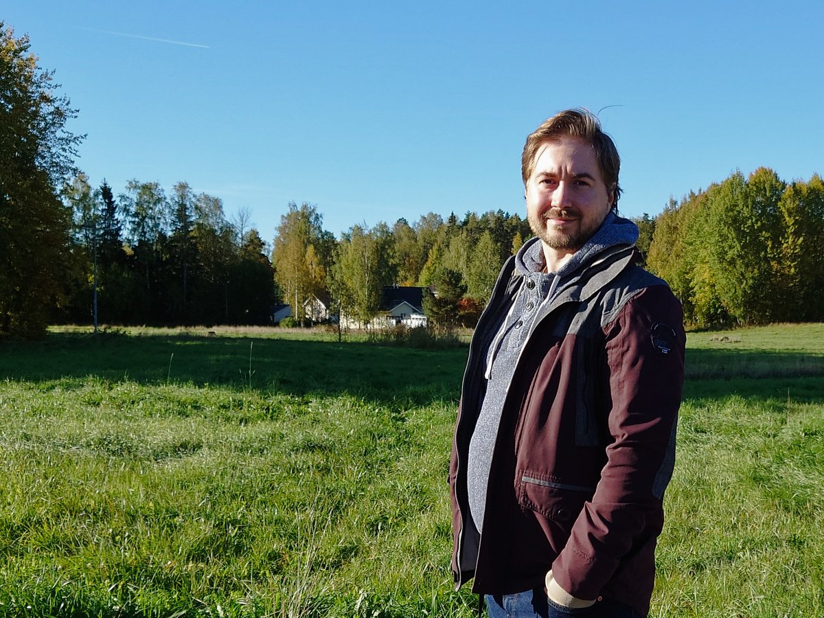 Lauri Lavanti in a dark jacket standing in a green field with autumn trees behind him.