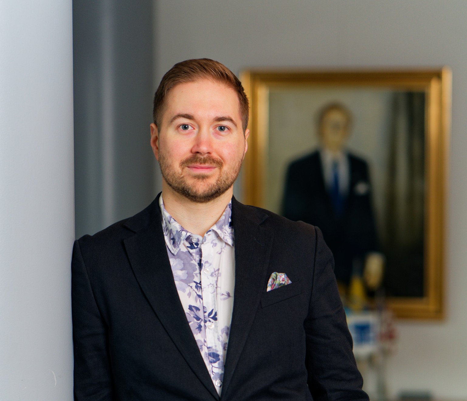 Lauri Lavanti leaning on a column with a portrait in the background, looking at the camera, smiling. He is wearing a white shirt with blue flower patterns and a dark jacket. Photo was taken by Juha Jantunen.
