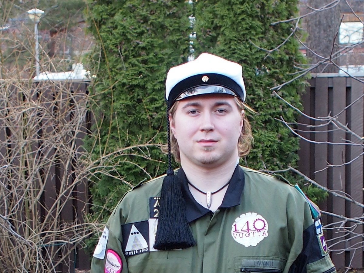 Lauri Lavanti in a tasselled student cap and a green student overall covered in patches, in a garden at Wappu.