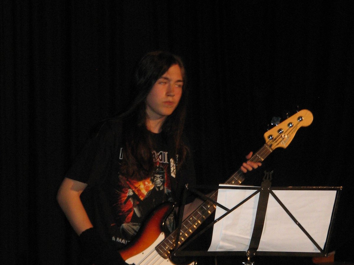A teenager with long hair playing a bass guitar alone on a dark stage, a music stand beside him.