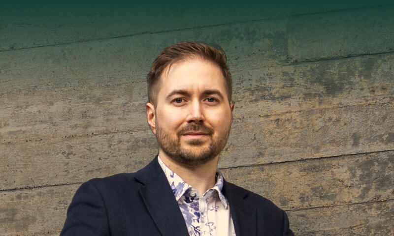Lauri Lavanti katsoo suoraan kameraan betoniseinän edessä, yllään tummansininen pikkutakki ja kukkakuvioinen paita. Kuva: Markus Isomeri.