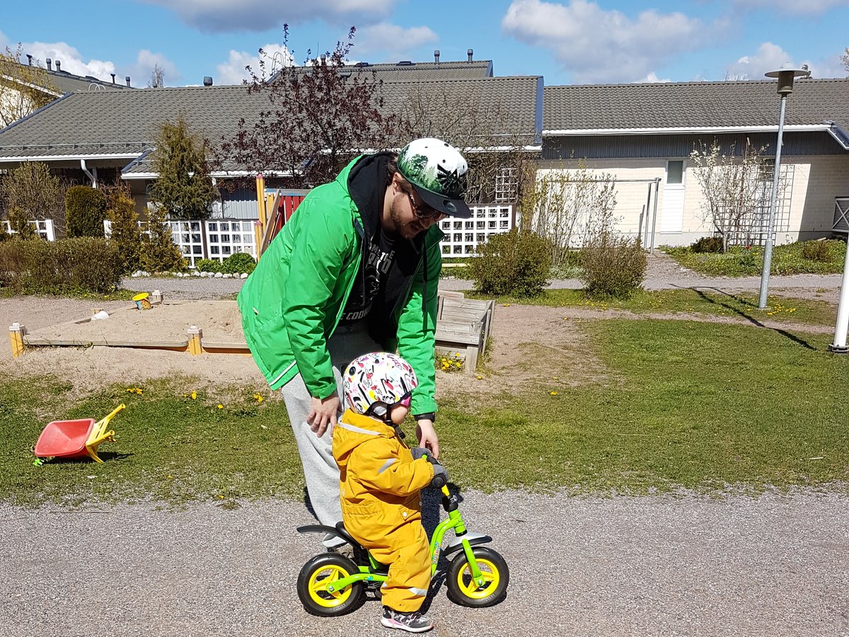 Lauri Lavanti in a green jacket bending over a small child in a yellow overall and helmet on a balance bike in a yard.