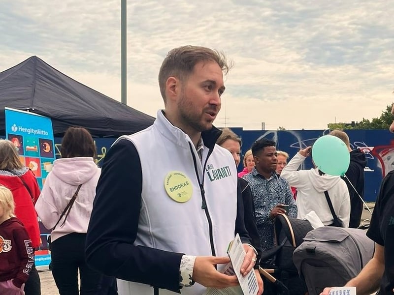 Lauri Lavanti in a white campaign vest with a candidate badge, handing out leaflets at a market square with people and a stroller behind him.