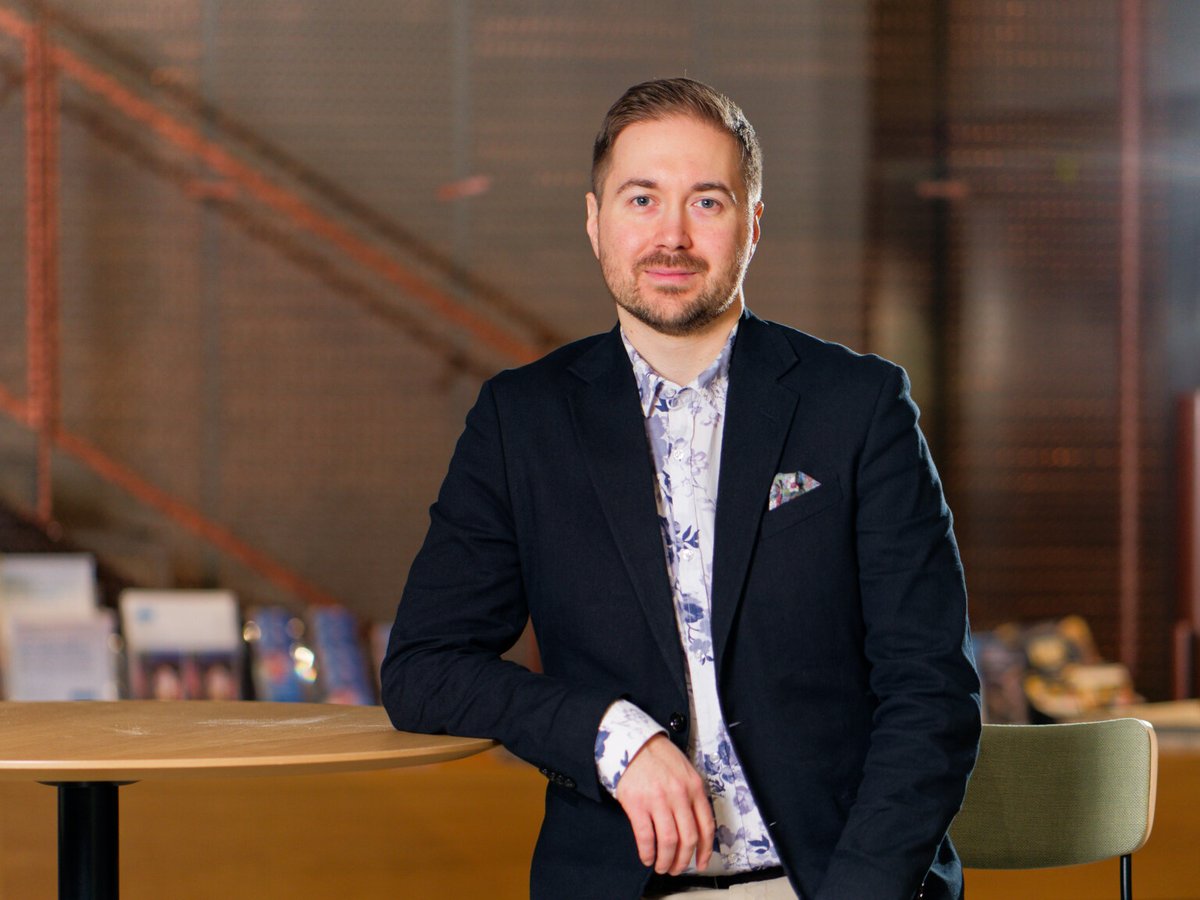 Lauri Lavanti in a navy blazer leaning on a high table, brochure shelves and a staircase behind him.