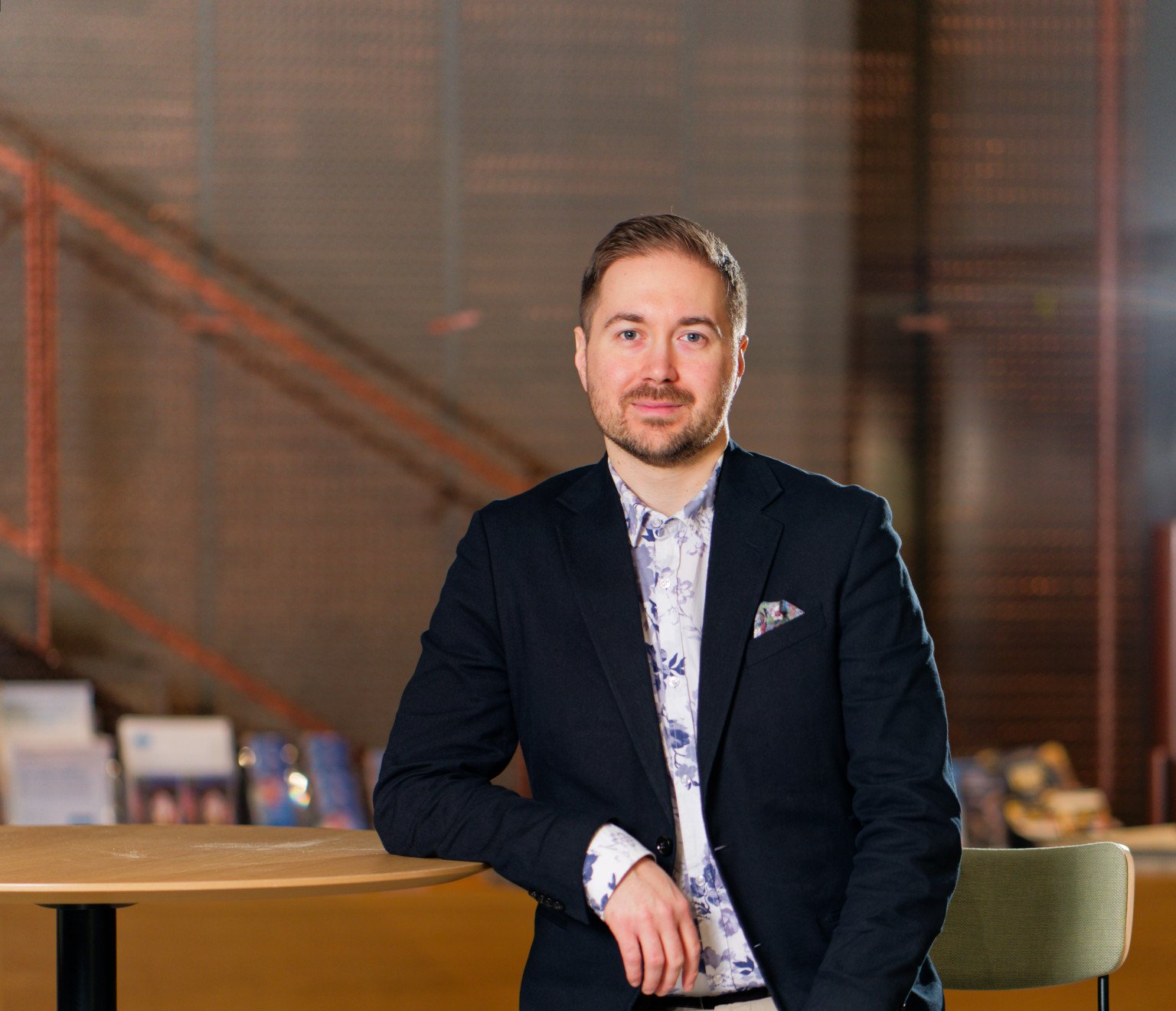 Lauri Lavanti sitting on a stool leaning against a tall table. He is wearing a black blazer and a blue-and-white floral shirt. In the blazer pocket is a multicoloured pocket square. A staircase in the background.