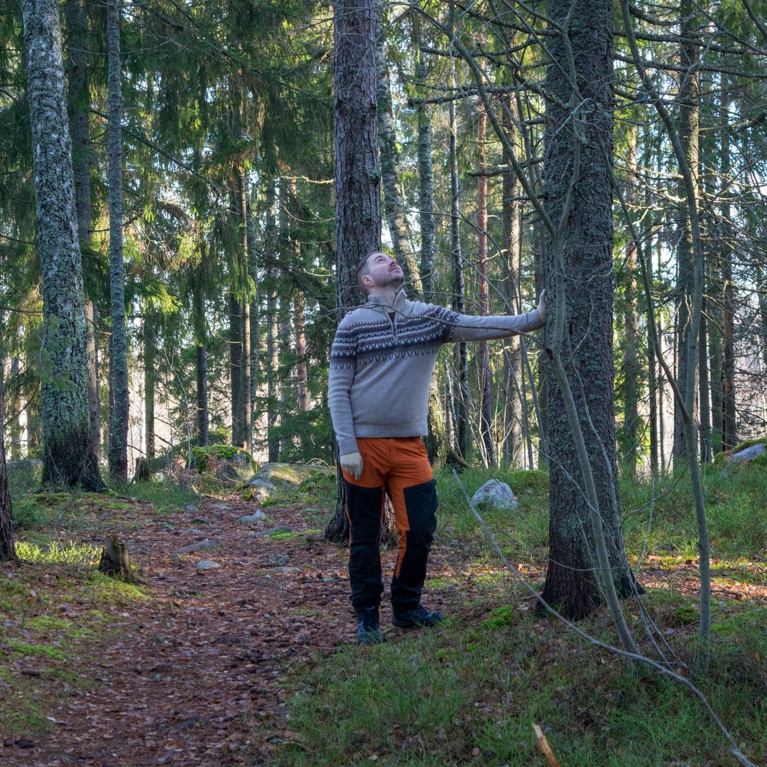 Lauri Lavanti in outdoor clothing in a forest, leaning against a tree and looking up its trunk.