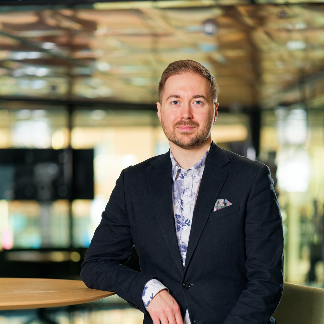 Lauri Lavanti leaning against a tall table. He is wearing a black blazer and a blue-and-white floral-patterned shirt. There is a multicoloured pocket square in the blazer pocket. Windows in the background.