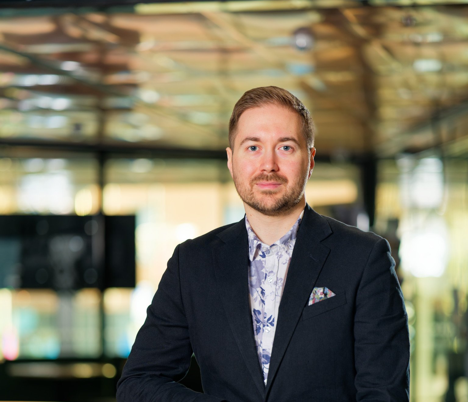 Lauri Lavanti sitting on a stool in front of a window, looking at the camera, smiling. He is wearing a white shirt with blue flower patterns and a dark jacket. Photo was taken by Juha Jantunen.