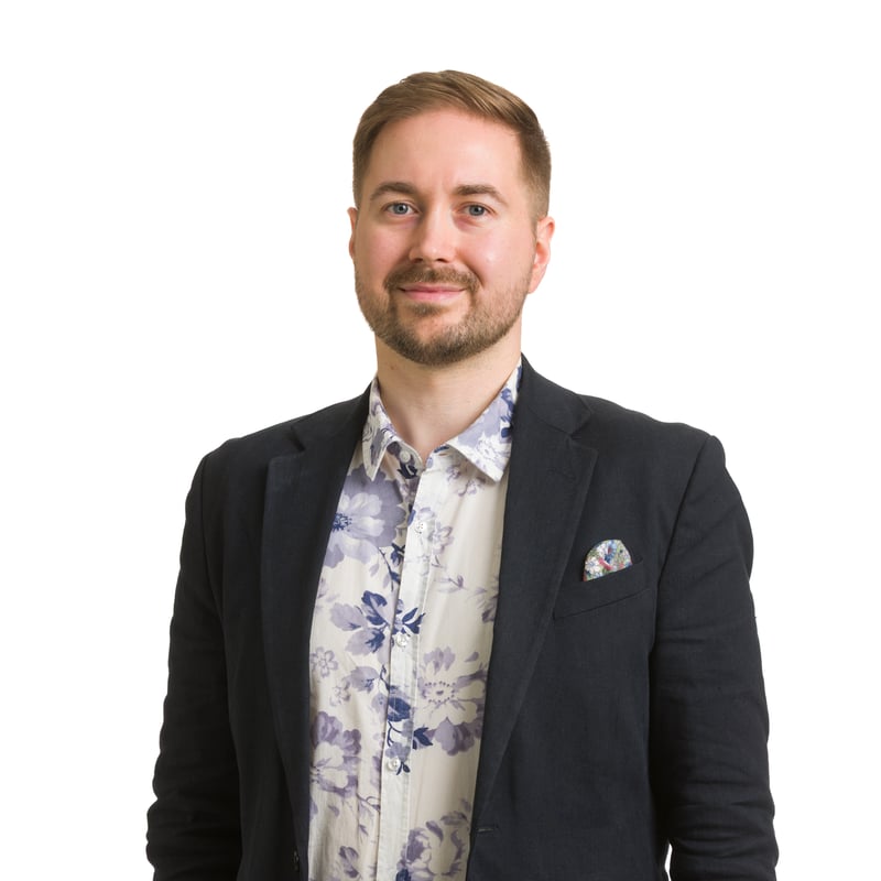 Studiokuva Lauri Lavannista valkoista taustaa vasten, toinen käsi taskussa, katse hieman sivulle, yllään tummansininen pikkutakki ja kukkakuvioinen paita.