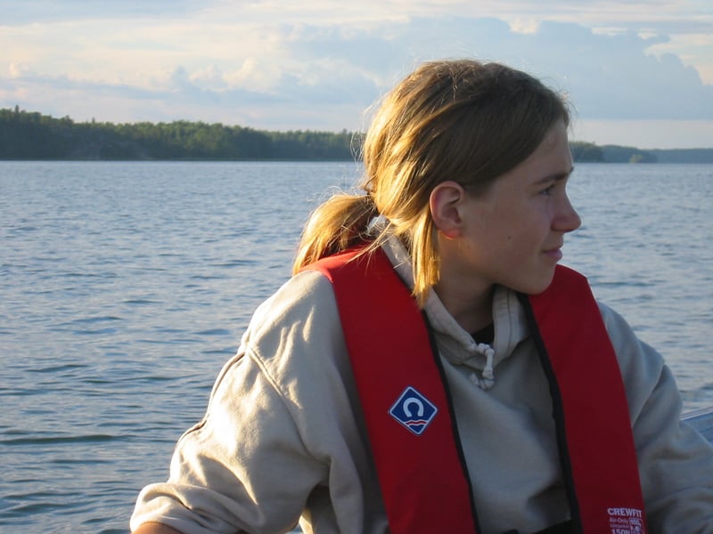A boy in a red life vest sitting in a boat, looking out over a calm sea with a wooded shore behind.