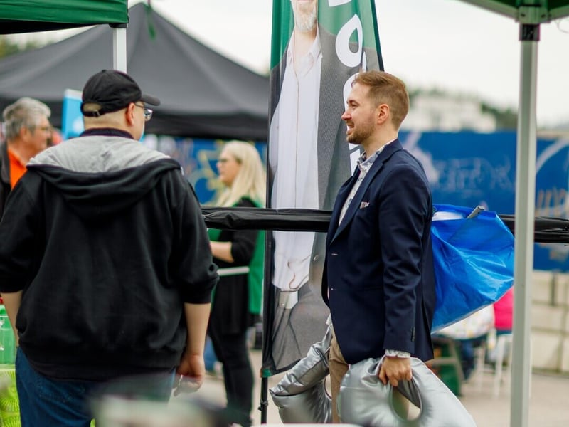 Lauri Lavanti in a navy blazer talking with a visitor at a green market tent, a campaign flag behind him.