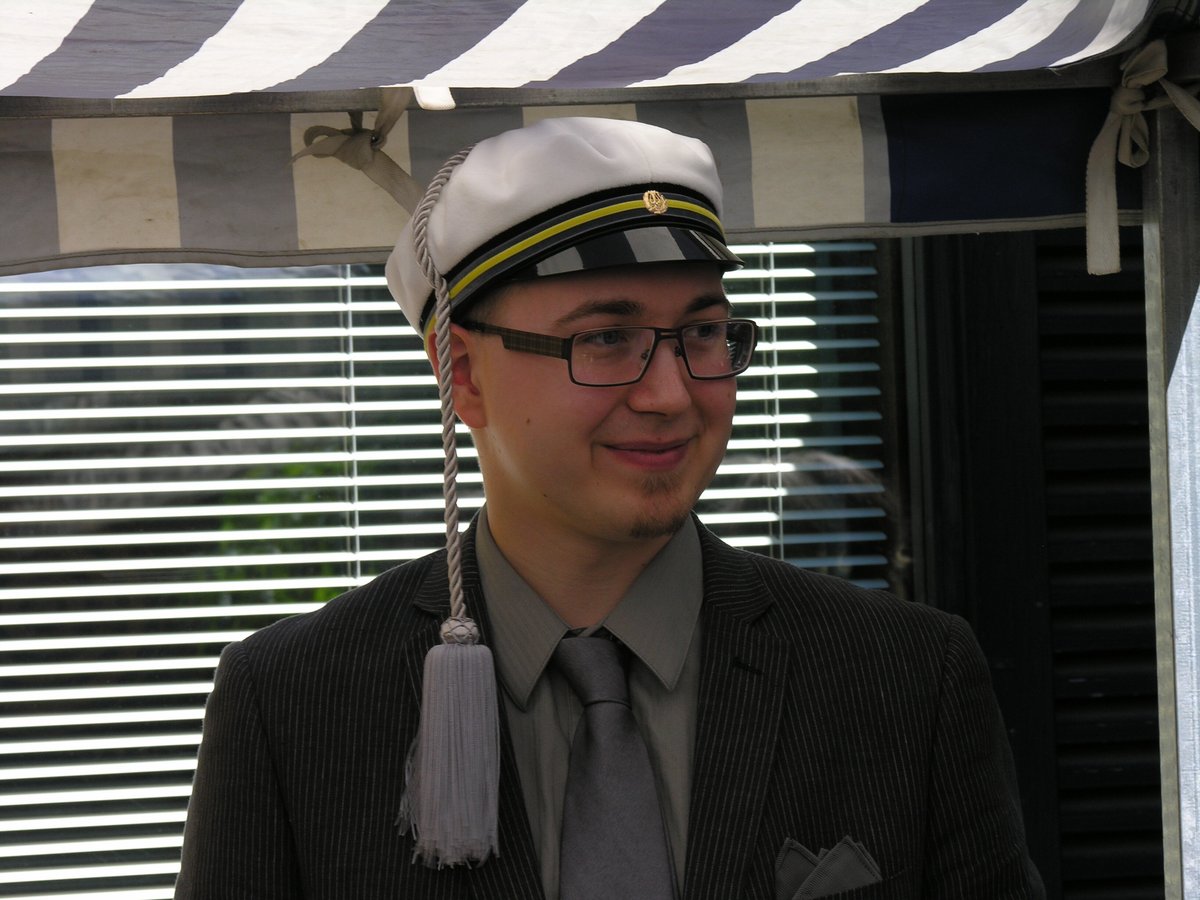 Lauri Lavanti in a white student cap and a grey suit, smiling under a striped awning on graduation day.