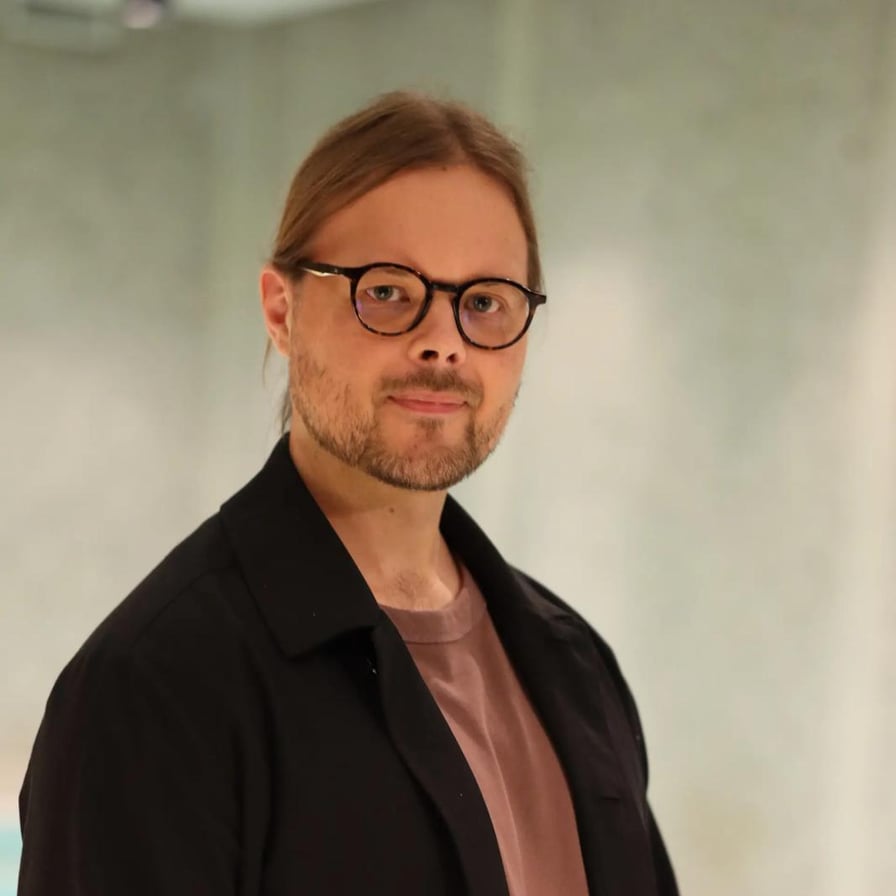 Kuva Lauri Nevanperästä. Hänellä on pitkät kammatut hiukset, parta ja silmälasit, päällään musta takki ja vaaleanpunertava t-paita.
