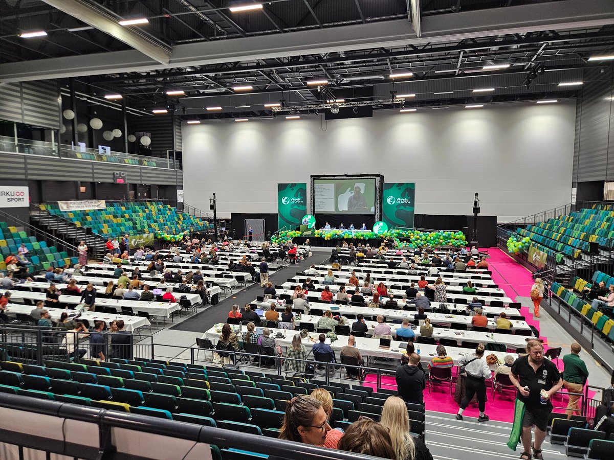 A sports hall set up as a party congress: rows of delegate tables, a stage with a large screen and green decorations.